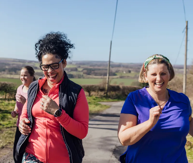 Three women power-walking and laughing together outdoors on a country lane.