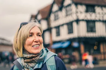 A smiling older woman out and about in a Warwickshire town, with a historic timber-framed building behind her.