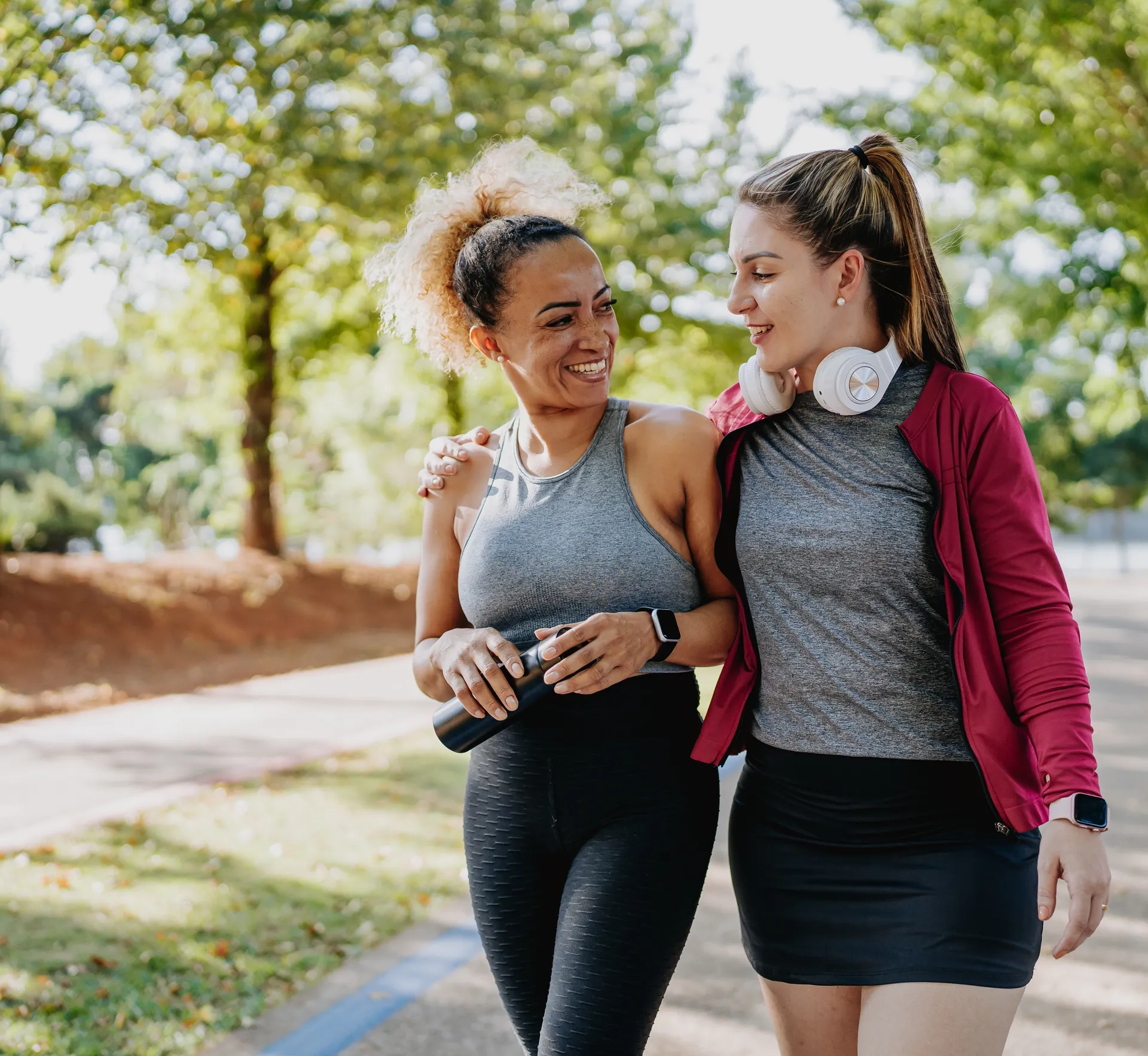 Two women in sportswear walking and chatting together along a park path.