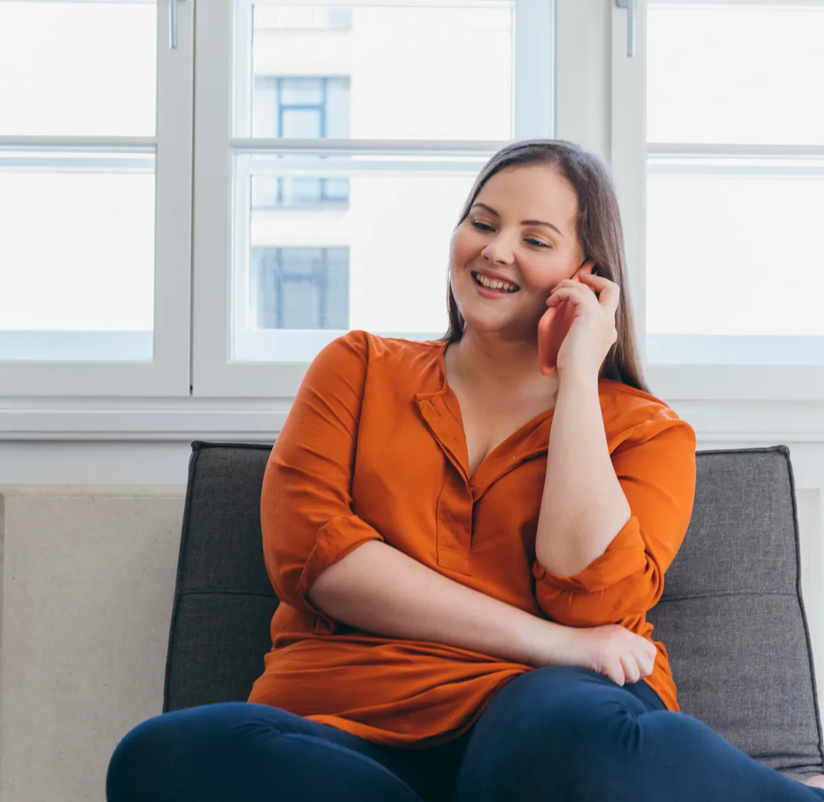 A woman sitting on her sofa by the window, smiling as she chats on the phone at home.