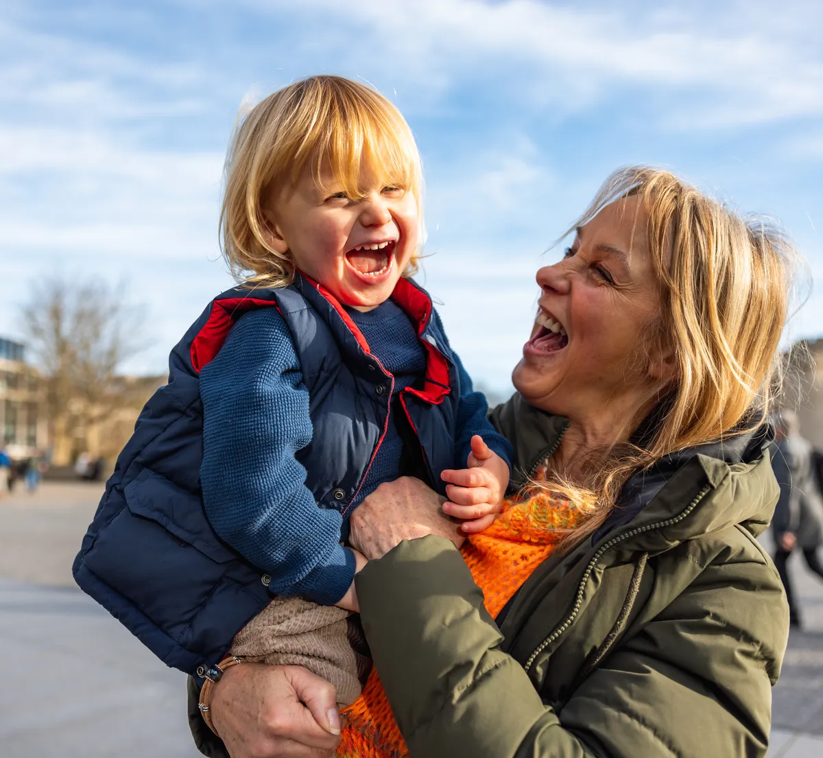 A grandmother laughing as she holds her young grandchild outdoors.
