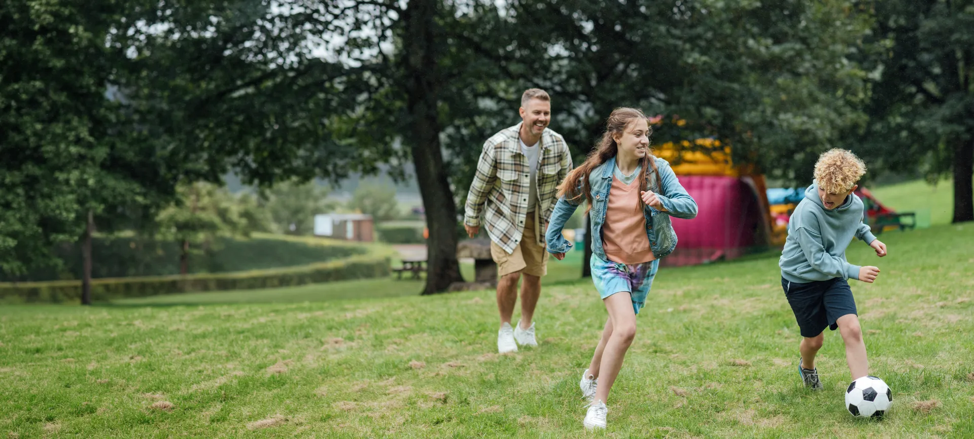 A father and his children laughing as they play football together in a local park.